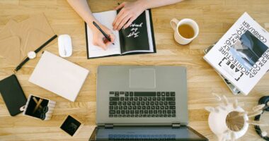 Woman writing notes at a home office desk with a laptop and coffee, representing planning a content calendar