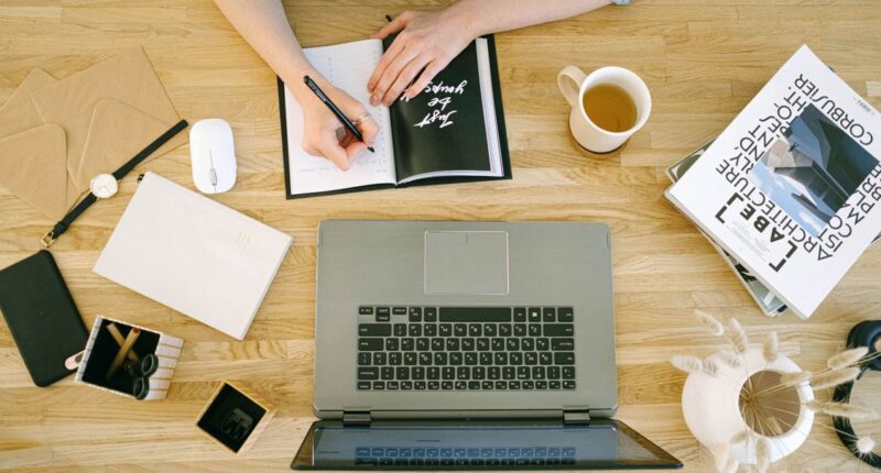 Woman writing notes at a home office desk with a laptop and coffee, representing planning a content calendar
