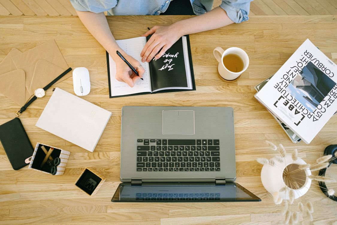 Woman writing notes at a home office desk with a laptop and coffee, representing planning a content calendar